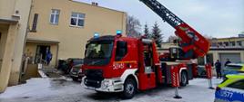 Czerwony wóz strażacki z wysuniętym długim podnośnikiem koszowym stoi na zaśnieżonym podwórzu przy budynku. Podnośnik sięga w górę wzdłuż ściany budynku. W tle widać inne budynki, drzewa oraz kilka osób, w tym funkcjonariuszy policji i zaparkowane radiowozy.