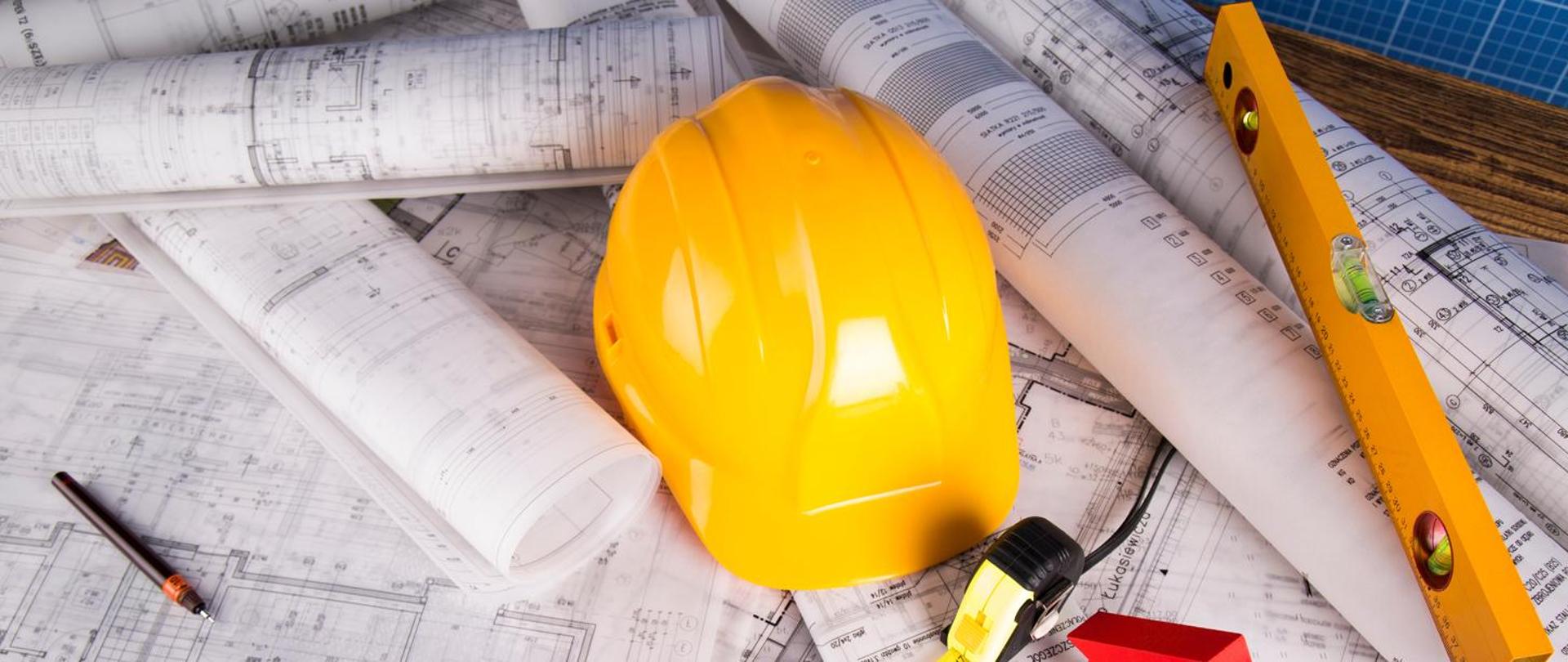 na środku leży żółty kask , obok metrówka, pomarańczowa poziomica, długopis. Wszystkie przedmioty leżą na rysunkach technicznych budynków.