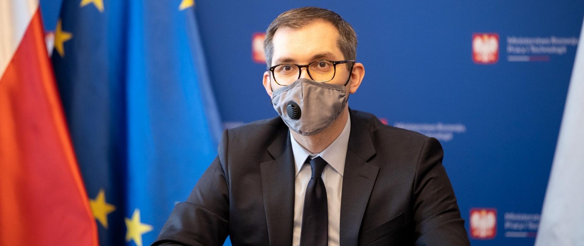 Wiceminister rozwoju, pracy i technologii Marek Niedużak siedzący za stołem, po jego lewej stroni flagi Polski i UE