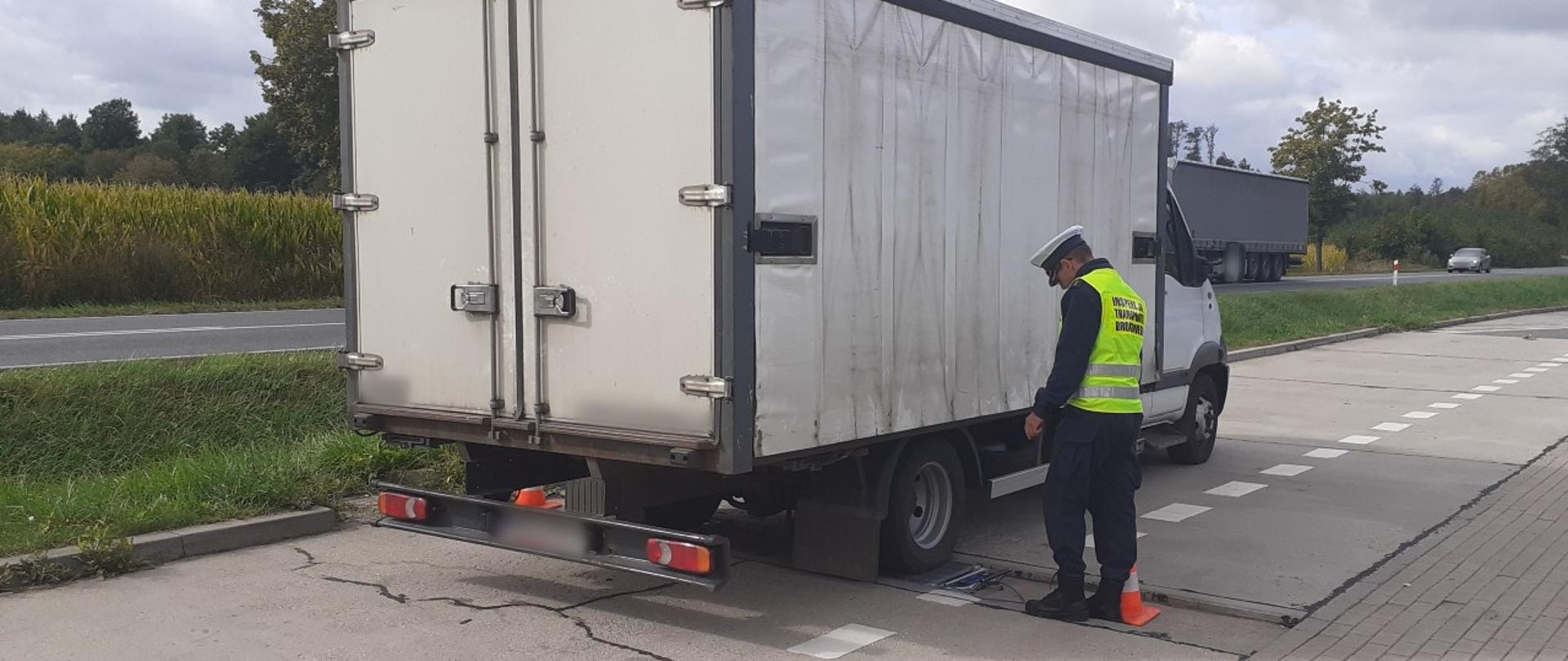 Umundurowany inspektor warmińsko-mazurskiej ITD stoi obok zatrzymanego samochodu dostawczego z napojami i odczytuje wyniki z wag, przez które przejeżdża bus.