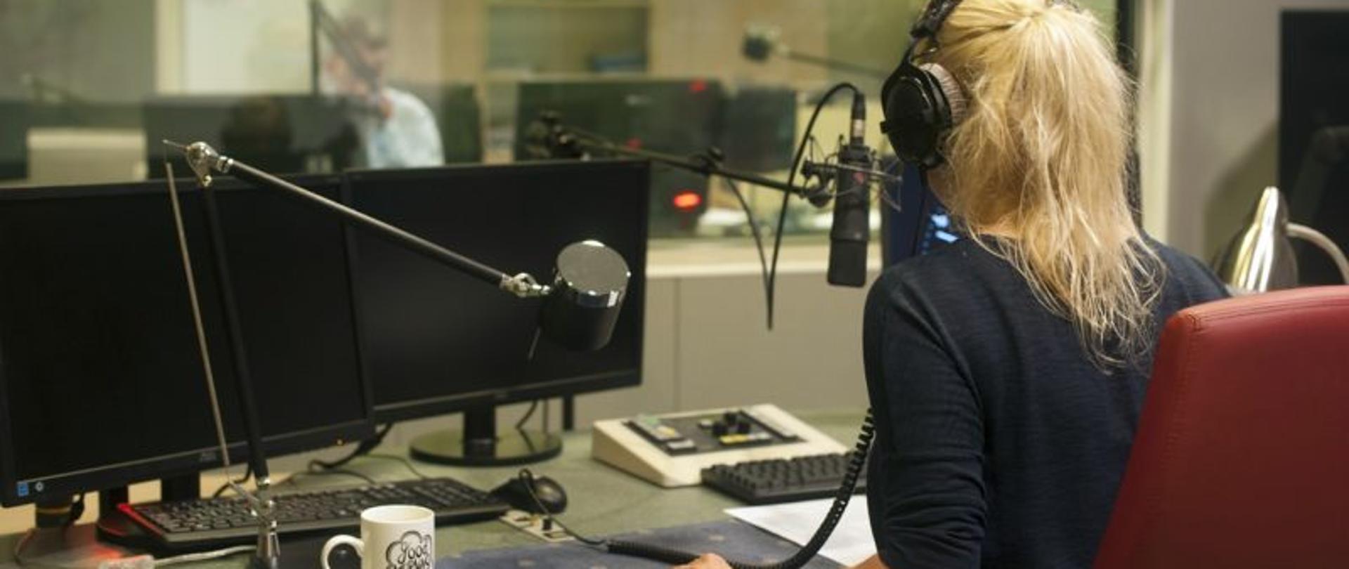Kobieta w studio radiowym , siedząca tyłem, za szybą, przed monitorem, ze słuchawkami na uszach.