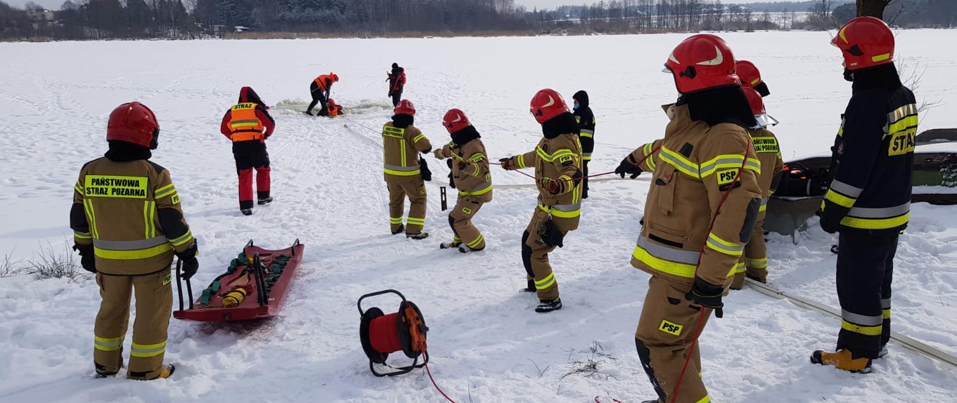 Na zdjęciu widać strażaków stojących na brzegu zamarzniętego zbiornika wodnego. Strażacy mają na sobie ubrania specjalne w kolorze żółtym i granatowym, na głowach czerwone hełmy. W oddali widać ratownika znajdującego się przy przerębli. W przerębli znajduje się kolejny strażak pozorujący osobę pod którą załamał się lód. Strażacy na brzegu naciągają i trzymają linę do której przywiązany jest ratownik niosący pomoc osobie znajdującej się w wodzie. Dokoła na ziemi leży śnieg. 