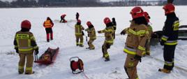 Na zdjęciu widać strażaków stojących na brzegu zamarzniętego zbiornika wodnego. Strażacy mają na sobie ubrania specjalne w kolorze żółtym i granatowym, na głowach czerwone hełmy. W oddali widać ratownika znajdującego się przy przerębli. W przerębli znajduje się kolejny strażak pozorujący osobę pod którą załamał się lód. Strażacy na brzegu naciągają i trzymają linę do której przywiązany jest ratownik niosący pomoc osobie znajdującej się w wodzie. Dokoła na ziemi leży śnieg. 
