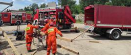 Ratownicy sgpr docinają na pile ukośnicy drewno potrzebne do stabilizacji budynku. Czynności wykonywane na zewnątrz budynku. Od lewej strony widać następujące pojazdy: specjalistyczny pojazd poszukiwawczo ratowniczy, następnie specjalistyczny ciężki pojazd ratownictwa technicznego oraz specjalistyczny ciężki pojazd kwatermistrzowski.