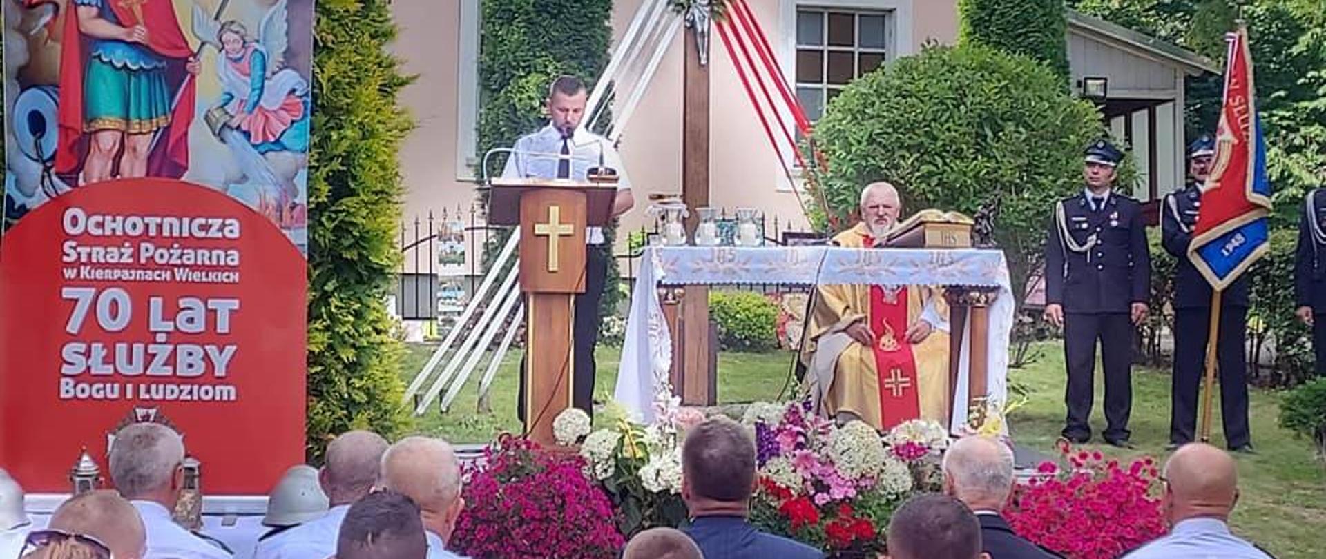 Na zdjęciu ołtarz na podwórku, w trakcie mszy, ludzie w ławkach z prawej strony sztandar strażacki. Z tyłu w oddali kościół.