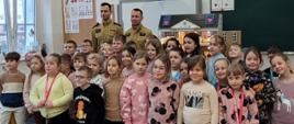 Zdjęcie przedstawia grupę dzieci z klas szkoły podstawowej. Wśród nich stoją funkcjonariusze w umundurowaniu służbowym. Zdjęcie jest wykonane w sali lekcyjnej