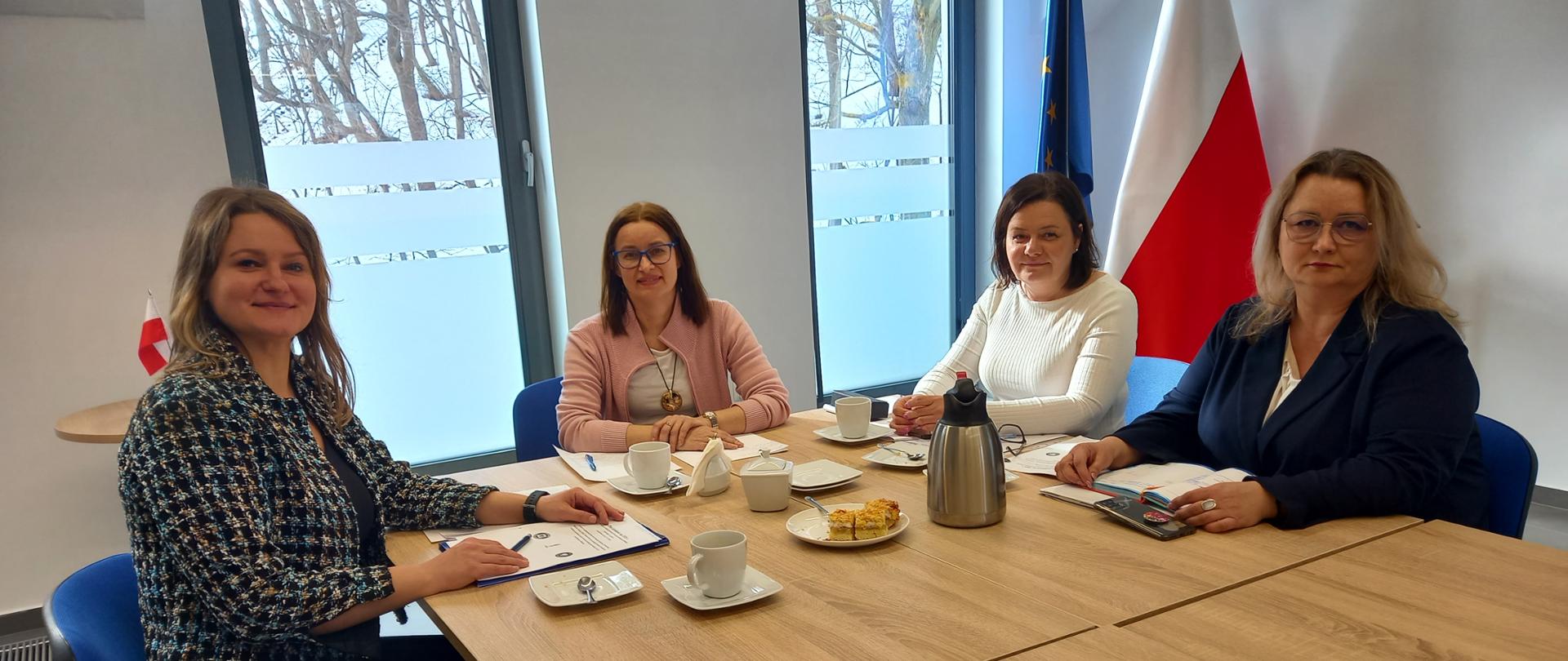 Cztery kobiety siedzące przy stole, za nimi dwie flagi: Polski i Unii Europejskiej, za oknami widać drzewa pozbawione liści i śnieg