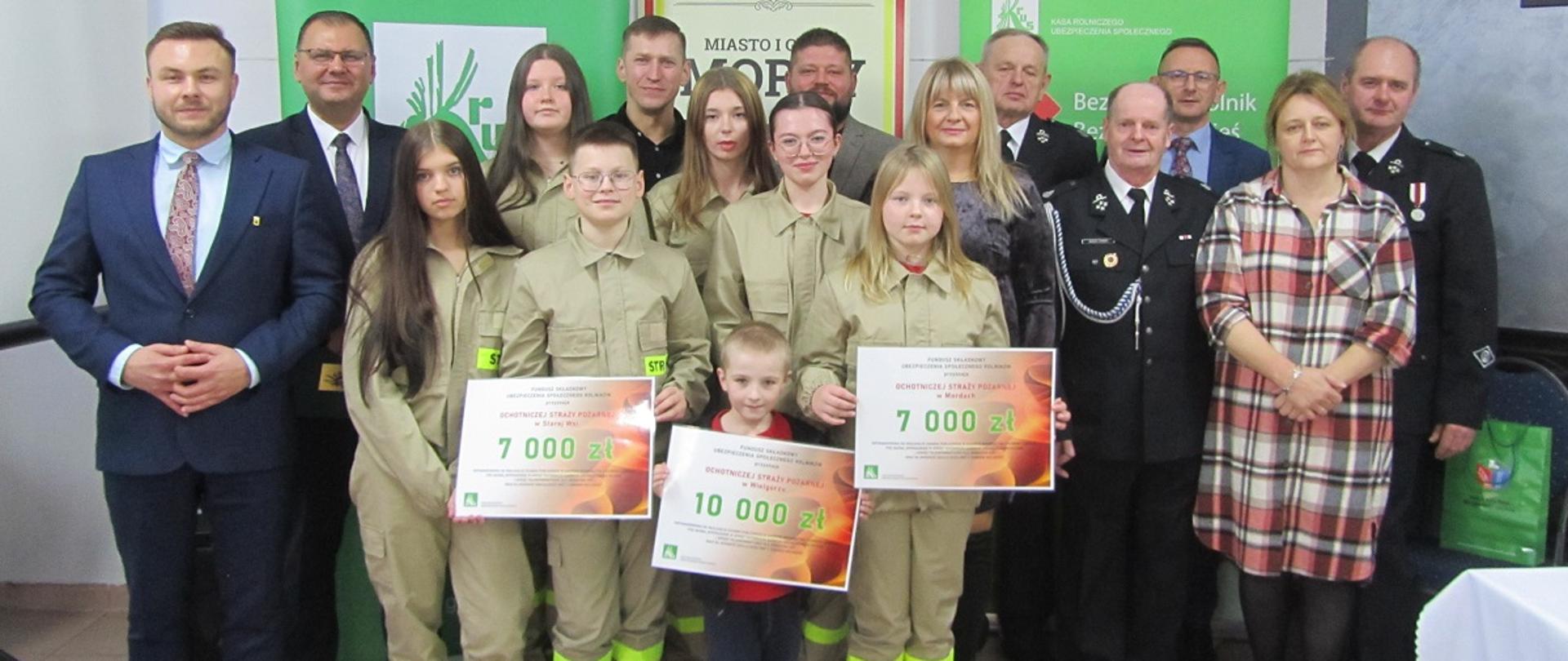 Grupa osób pozuje do zdjęcia w pomieszczeniu. Za nimi stoją trzy banery. Dwa z logiem KRUS i jeden z napisem Miasto i Gmina Mordy. W pierwszym rzędzie stoją dzieci i młodzież w jasnozielonych strojach z napisem „STRAŻ” trzymają duże symboliczne vouchery. Za nimi stoją dorośli- część w eleganckich garniturach, część w mundurach strażackich oraz cywilnych ubraniach. Wszyscy uśmiechają się w stronę aparatu, co sugeruje uroczyste wręczenie voucherów.