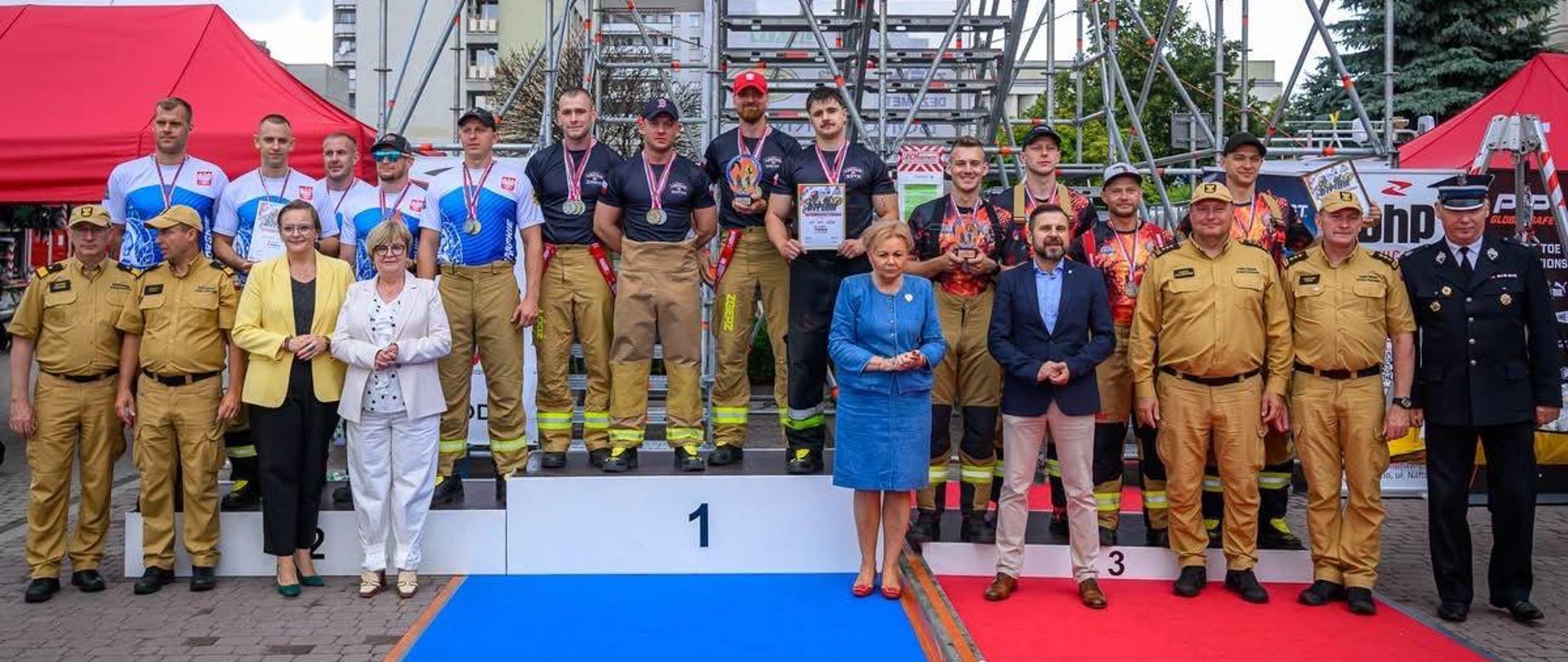 Na trzystopniowym podium stoją zawodnicy w pełnym umundurowaniu sportowym strażaka: bojowe spodnie, koszulki z motywem płomieni, medale na szyjach. Zespół zajmuje miejsce na podium, otoczony przez organizatorów i przedstawicieli służb w galowych mundurach. Za nimi wznosi się wysoka konstrukcja rusztowania, a po bokach stoją czerwone namioty. Zdjęcie przedstawia oficjalny moment dekoracji medalistów — brązowych zdobywców z KW PSP Opole.