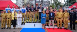Na trzystopniowym podium stoją zawodnicy w pełnym umundurowaniu sportowym strażaka: bojowe spodnie, koszulki z motywem płomieni, medale na szyjach. Zespół zajmuje miejsce na podium, otoczony przez organizatorów i przedstawicieli służb w galowych mundurach. Za nimi wznosi się wysoka konstrukcja rusztowania, a po bokach stoją czerwone namioty. Zdjęcie przedstawia oficjalny moment dekoracji medalistów — brązowych zdobywców z KW PSP Opole.