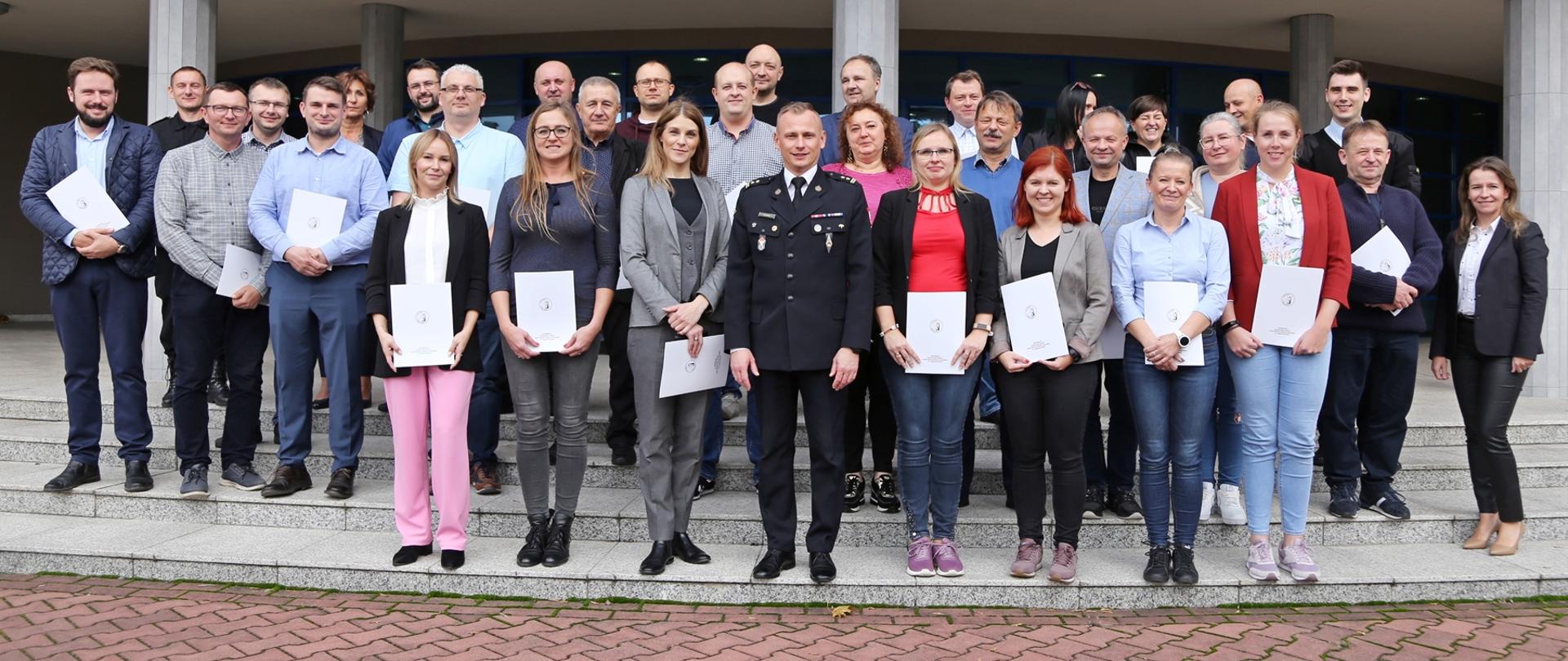 Grupowe zdjęcie uczestników szkolenia (29 osób) w towarzystwie Zastępcy Komendanta Centralnej Szkoły PSP, dwóch funkcjonariuszy CS PSP oraz kobiety