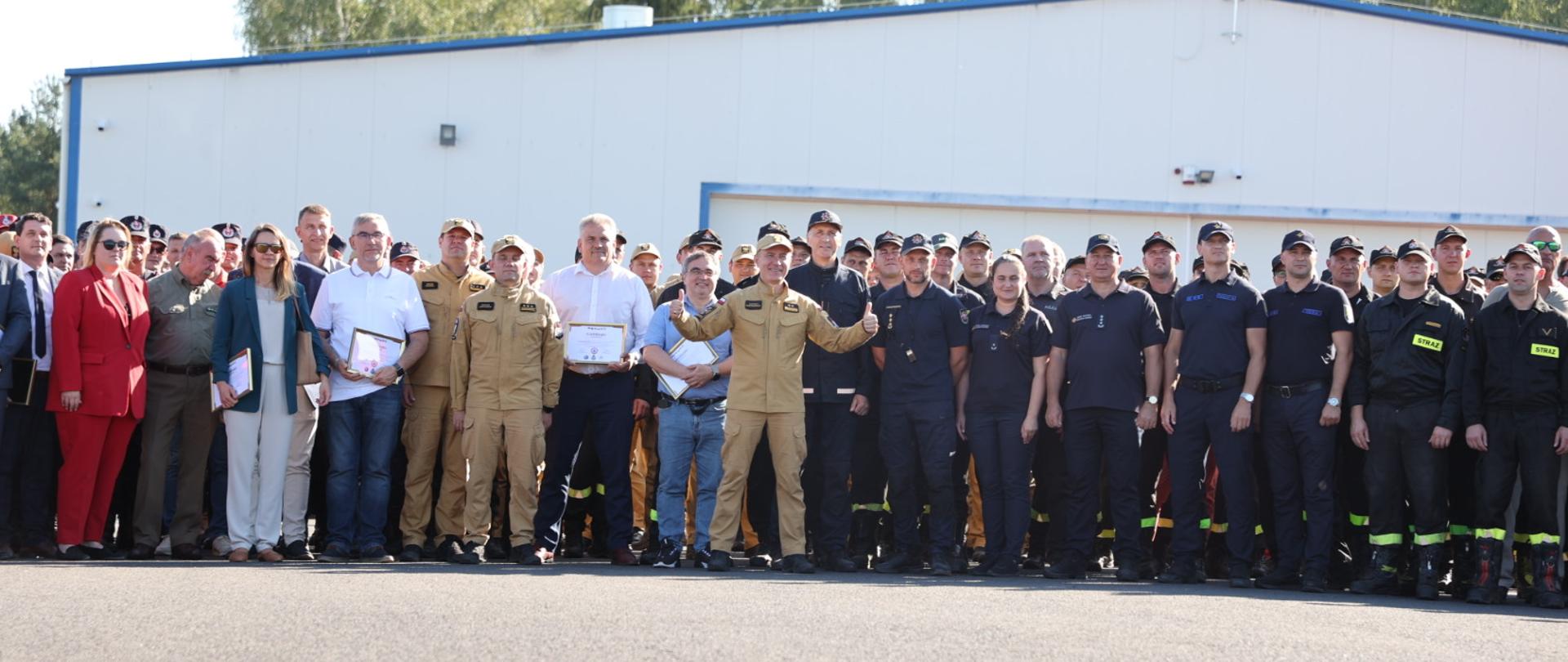 Na palcu komendant główny PSP stoi w grupie z uczestnikami i organizatorami ćwiczeń, komendant unosi obie ręce do góry z kciukiem w górę