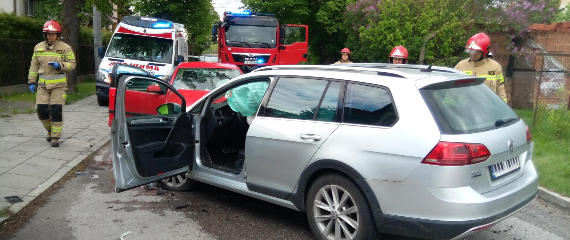 Zdjęcie przedstawia ulicę Skłodowskiej-Curie i stojące na niej zniszczone samochody. Za nimi znajdują się: po lewej stronie ambulans a po prawej samochód pożarniczy. Przy autach przechodzą strażacy. 