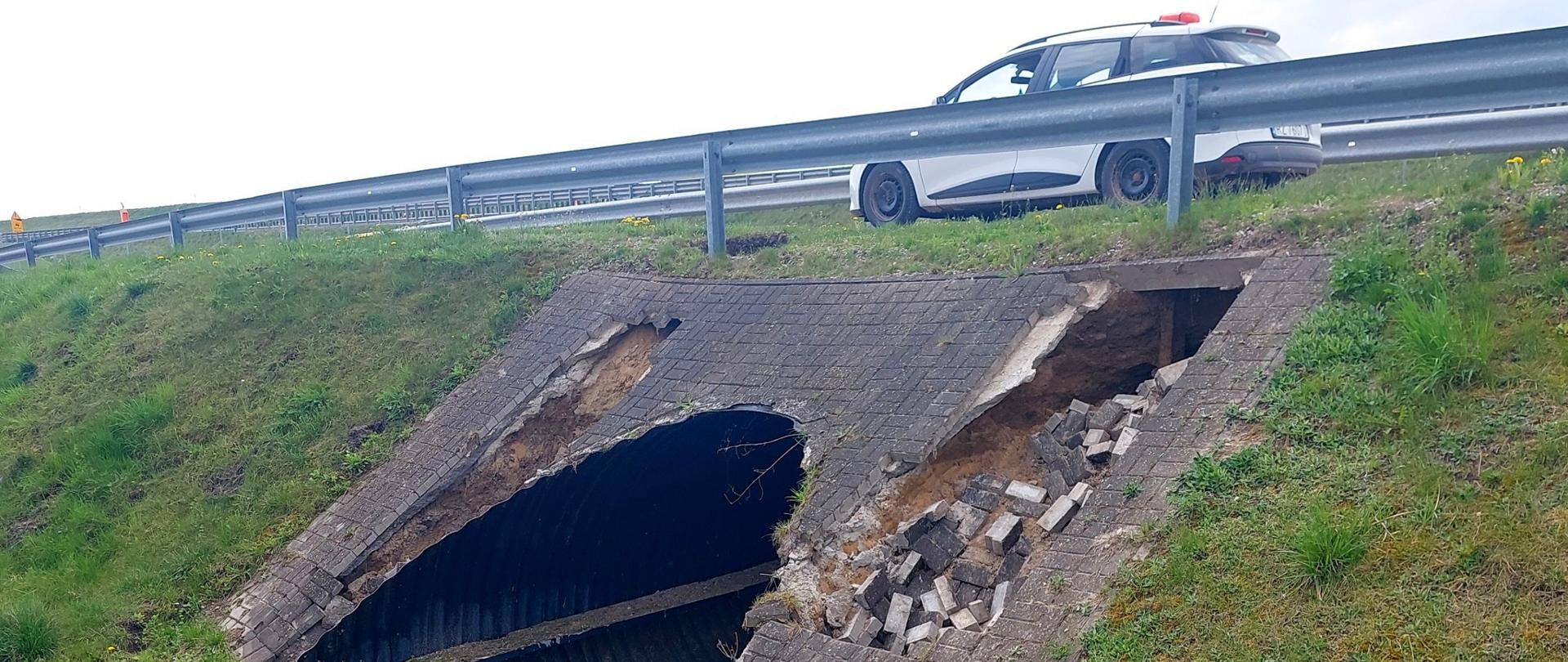 Widok na uszkodzony przepust po nawalnym deszczu. Wypłukana jest kostka, widać pusta przestrzeń w miejscu bariery drogowej. Na drodze stoi auto służby drogowej. 