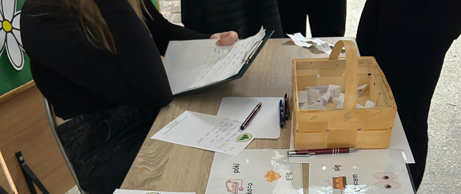 Stół z pomocami dydaktycznymi: koszyk z losami, czyste arkusze odpowiedzi oraz cztery zalaminowane karty z rebusami obrazkowymi wspomagającymi naukę ortografii i frazeologii.