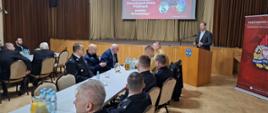 Strażacy ubrani w granatowe mundury galowe, oraz pisakowe mundury siedzą przy stolikach, w tyle rolap z logiem Państwowej Straży Pożarnej na tylnej ścianie kotary oraz ekran z wyświetloną prezentacją. Przy mównicy stoi wójt ubrany w garnitur, przed sobą ma mikrofon