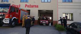 Plac przed komendą. Na tle budynku komendy stoi wóz strażacki, przed nim w dwuszeregu - strażacy w umundurowaniu służbowym w kolorze piaskowym oraz koszarowym. Na przeciwko nich znajduje się biało-czerwony maszt flagowy, przy którym stoi poczet flagowy w umundurowaniu wyjściowym. Flagowy podnosi flagę, dwaj pozostali członkowie pocztu salutują. Przodem do budynku stoi zastępca komendanta powiatowego w mundurze wyjściowym, ktory także oddaje honor przez salutowanie.
