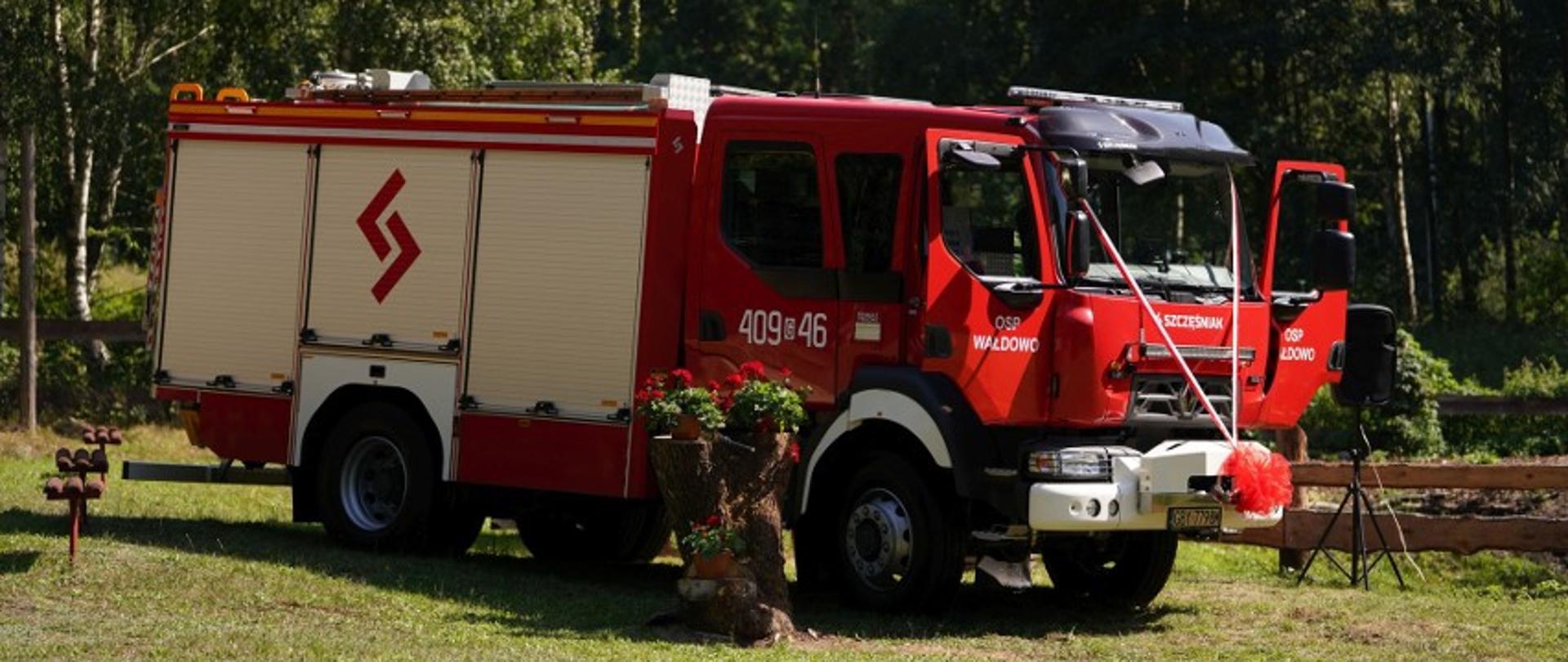 Zdjęcie przedstawia stojący na polanie wóz strażacki. Na masce ma on 2 wstęgi biało-czerwone, które łączą się na zderzaku. Drzwi do wozu są otwarte. Obok niego poustawiane są czerwone kwiaty.