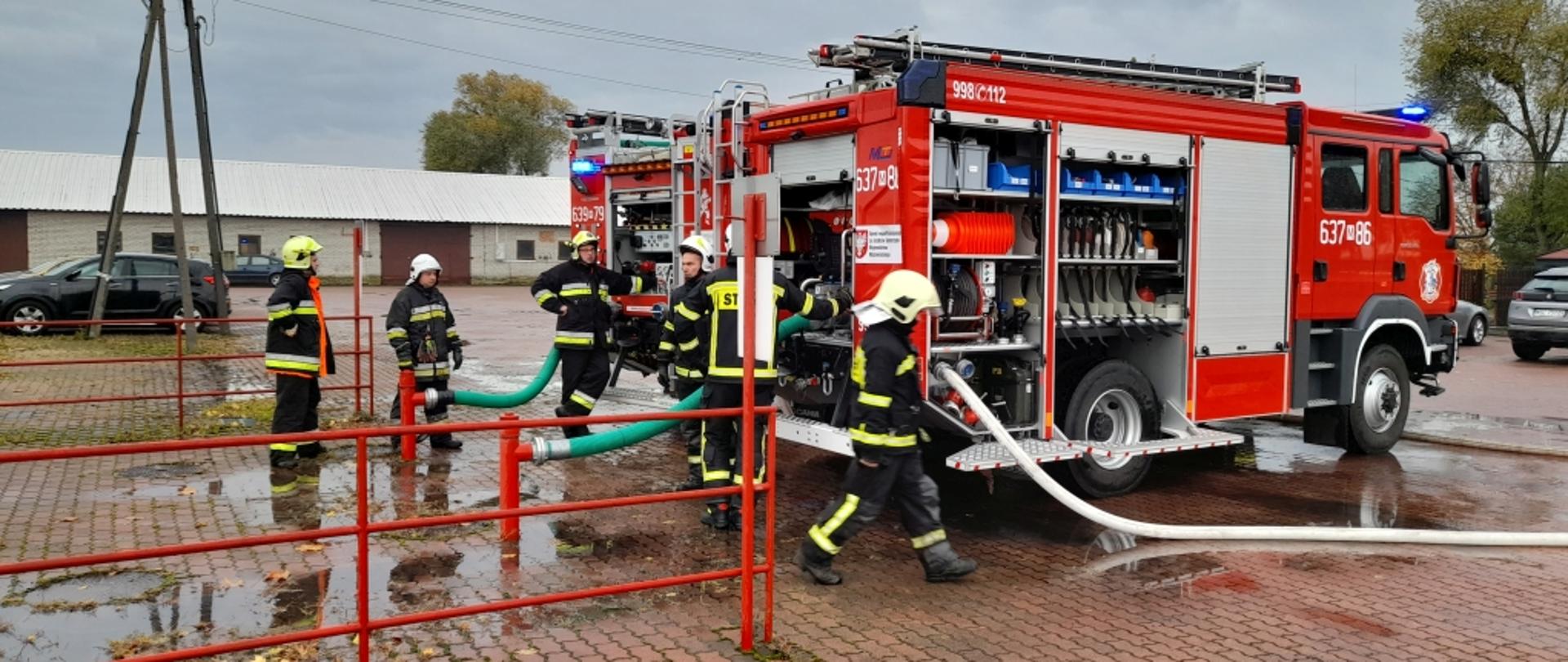 Na zdjęciu widoczne dwa samochody gaśnicze oraz kilku strażaków. Samochody zasysają wodę ze zbiornika przeciwpożarowego.