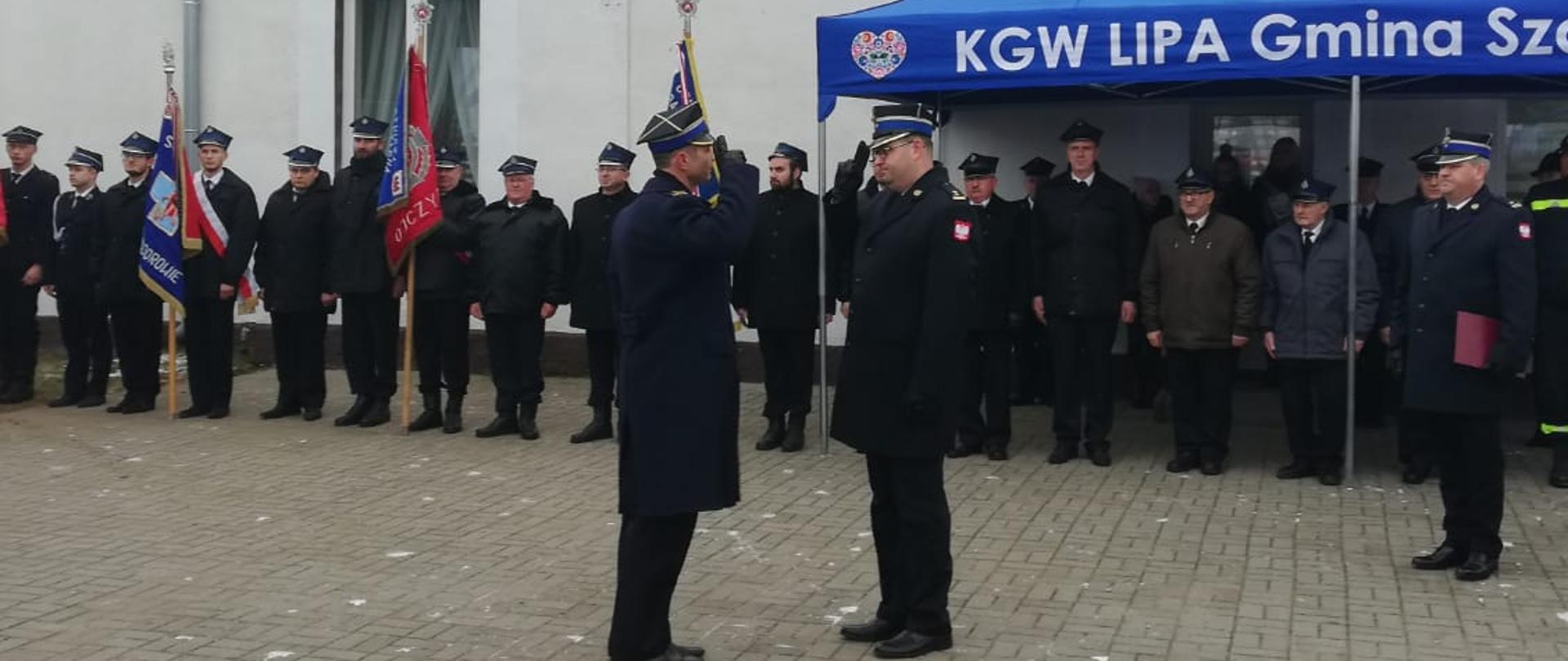 Składanie meldunku strażakowi przez drugiego strażaka. w tle poczty sztandarowe oraz budynek