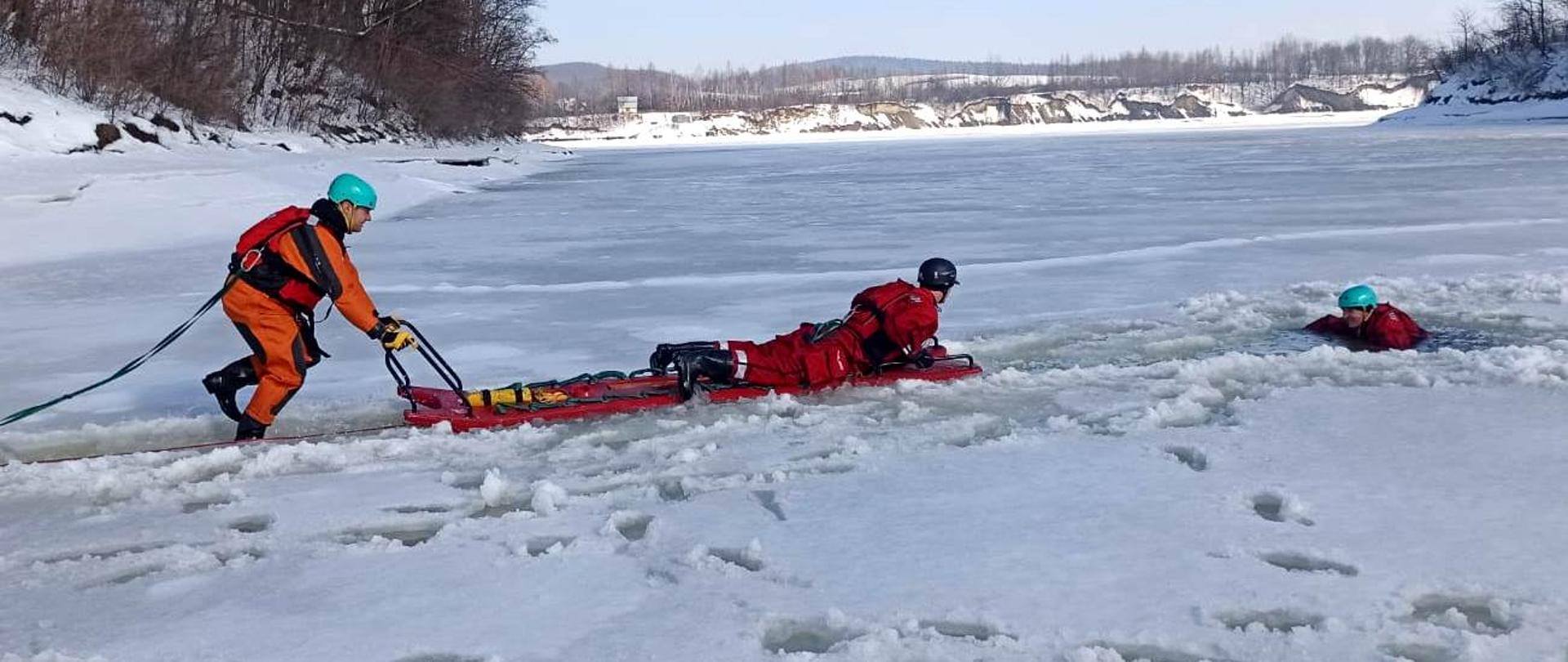 Na zdjęciu widocznych jest trzech strażaków ubranych w specjalne ubrania do pracy w wodzie oraz kaski ochronne, ćwiczących z zakresu ratownictwa lodowego. Strażacy znajdują się na zamarzniętym zbiorniku wodnym. Jeden z nich przebywa w wodzie w przerębli, dwóch pozostałych podejmuje próbę wyciągnięcia go z wody przy użyciu specjalnych sani lodowych.