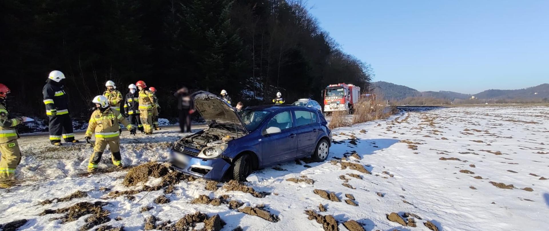 Samochód osobowy stoi na kołach poza drogą na zaśnieżonym polu. Wokół stoją strażacy. W tle stoi samochód pożarniczy.