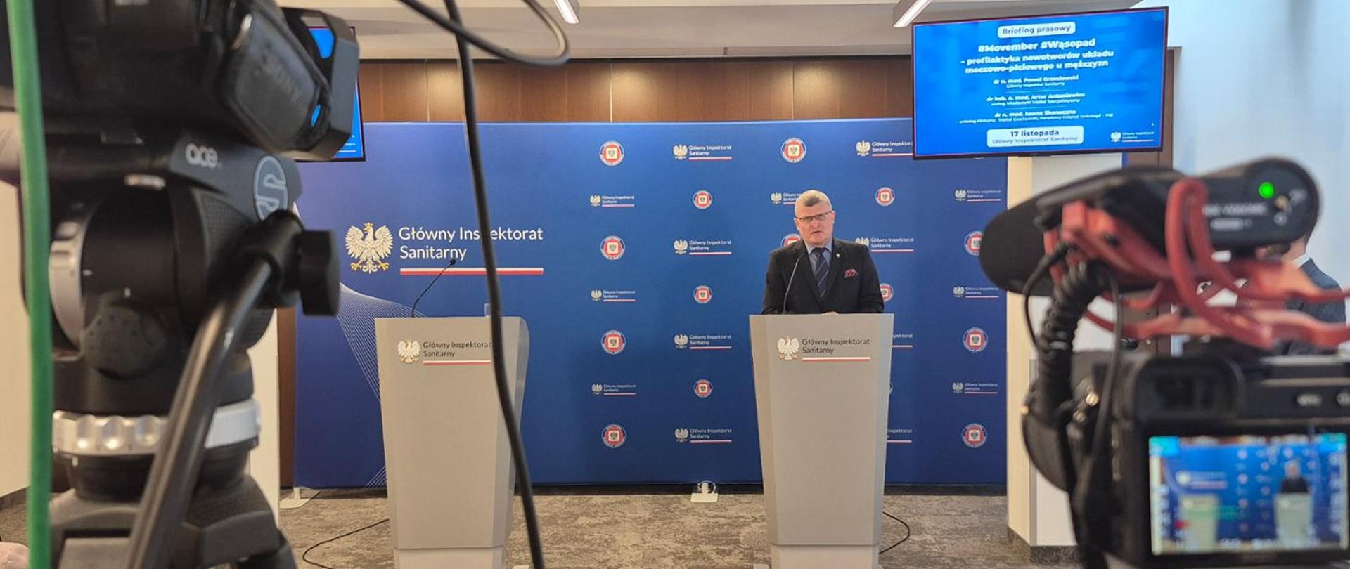 Na zdjęciu widać scenę z konferencji prasowej organizowanej przez Główny Inspektorat Sanitarny. W centrum stoi mężczyzna w garniturze za mównicą, przemawiający do kamer. Za nim znajduje się ściana z oficjalną grafiką GIS: godłem Polski, logotypami instytucji oraz napisem „Główny Inspektorat Sanitarny”.
Po obu stronach kadru widoczne są kamery nagrywające wydarzenie. Nad mównicą, z prawej strony, wisi ekran prezentujący slajd dotyczący profilaktyki nowotworów układu moczowo-płciowego u mężczyzn w ramach akcji Movember / Wąsopad.
Scena wygląda jak formalny briefing prasowy poświęcony zagadnieniom zdrowia publicznego.