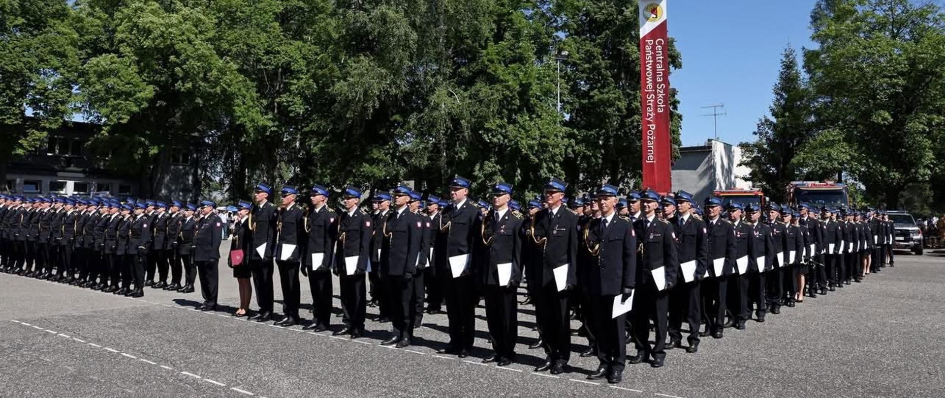 Zdjęcie przedstawia pododdział strażaków PSP ustawiony w równych szeregach na placu apelowym. Funkcjonariusze ubrani są w czarne mundury wyjściowe i czapki służbowe. W tle widoczna jest wieża szkoleniowa oraz drzewa otaczające plac. Pogoda jest słoneczna i bezdeszczowa.