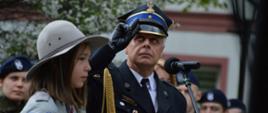 Narodowe Święto Trzeciego Maja. Plac Wolności w Rawiczu. Strażak - zastępca komendanta powiatowego PSP salutuje w czasie hymnu i podnoszenia flagi państwowej. Z jego lewej strony stoi harcerka w kapeluszu. W tle klasa mundurowa.