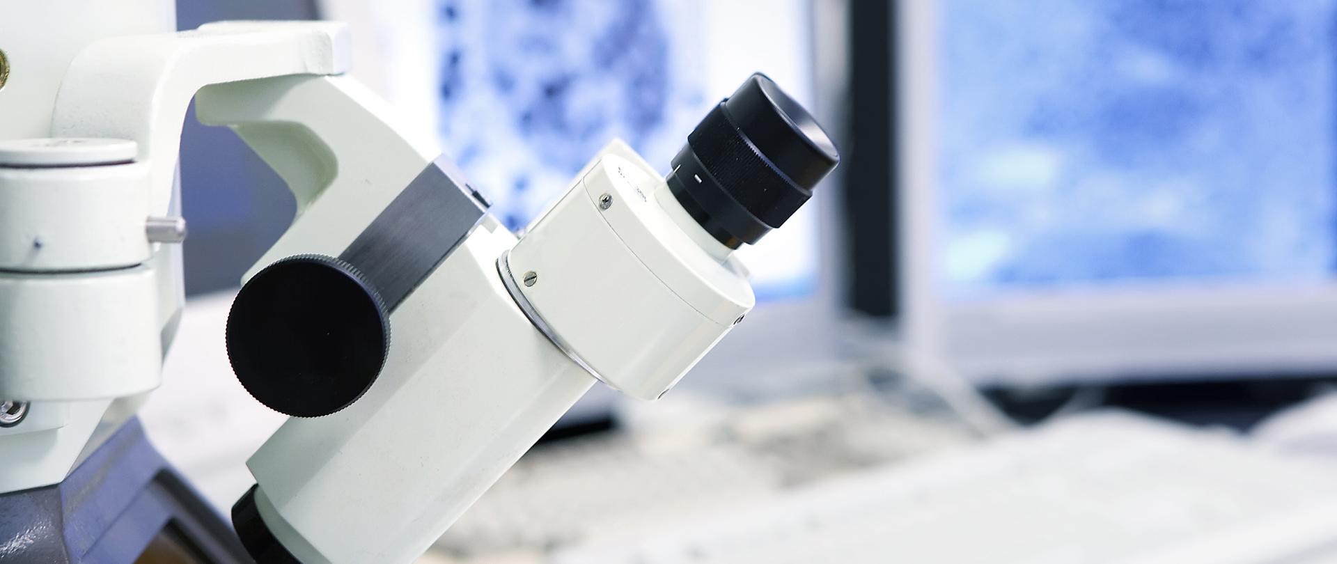 Na zdjęciu widać nowoczesny mikroskop, stojący na biurku. Za nim jest komputer.