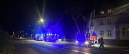 Widok na ulicę, na której rozstawione są pojazdy strażaków. Jest noc. Przy jednym z budynków przy jego ścianie stoi rozstawiony podnośnik. Zdjęcie oświetlone niebieskimi błyskami.