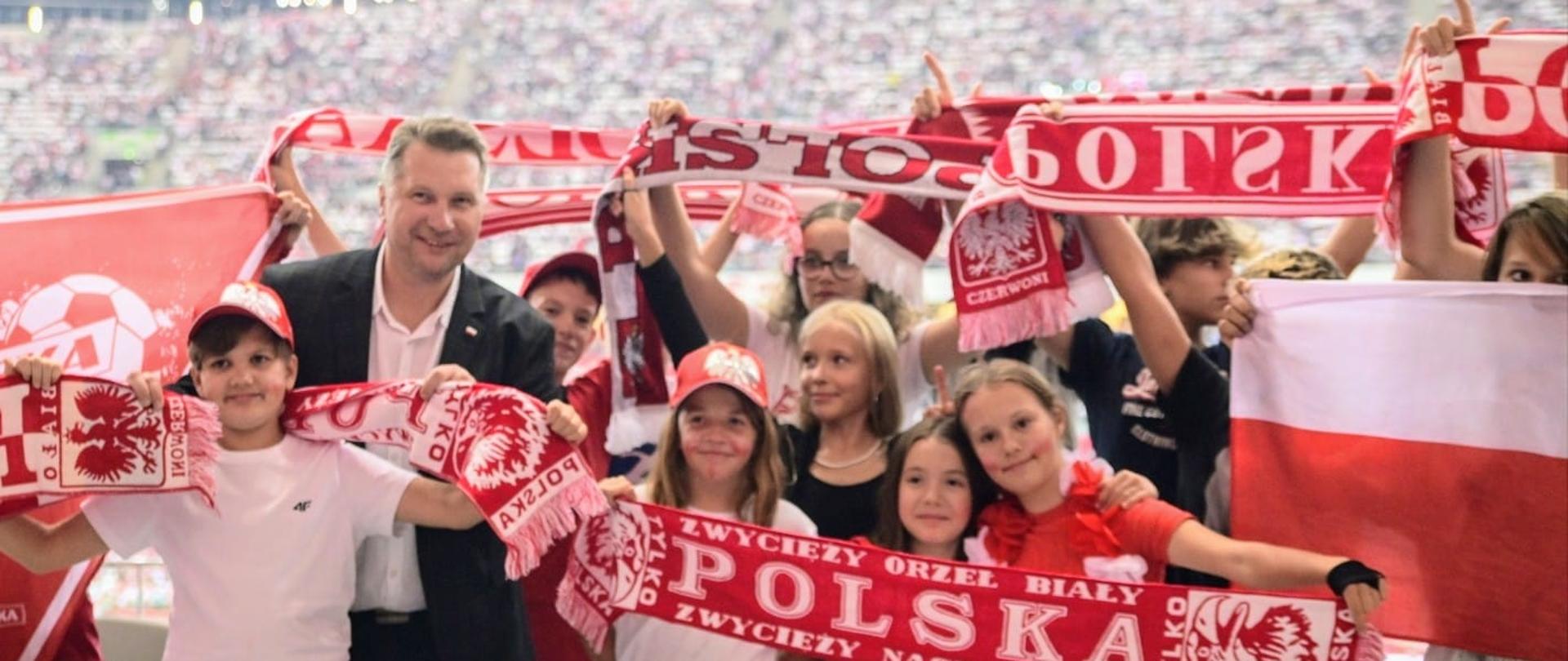 Minister Czarnek stoi wśród trzymających rozpostarte biało-czerwone szaliki dzieci, w tle wypełnione ludźmi trybuny stadionu.
