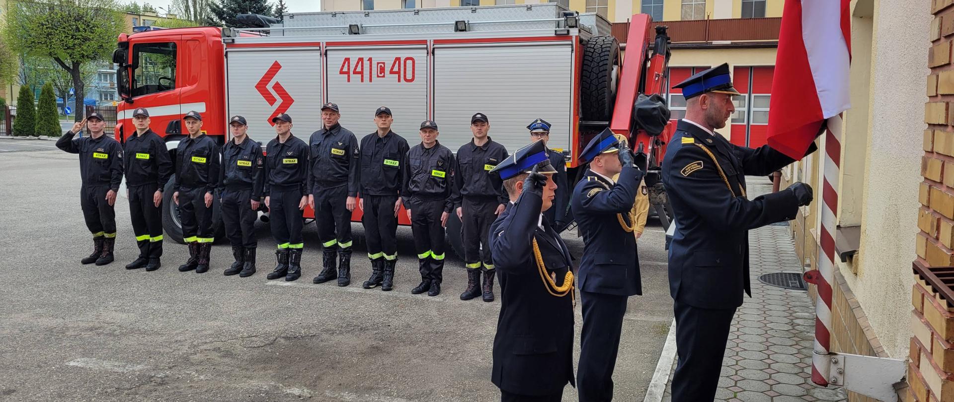 Strażacy w czarnych mundurach stojący w szeregu na utwardzonym placu. Na pierwszym planie jeden z nich wciąga biało-czerwoną flagę na maszt. W tle czerwono-srebrny strażacki samochód.