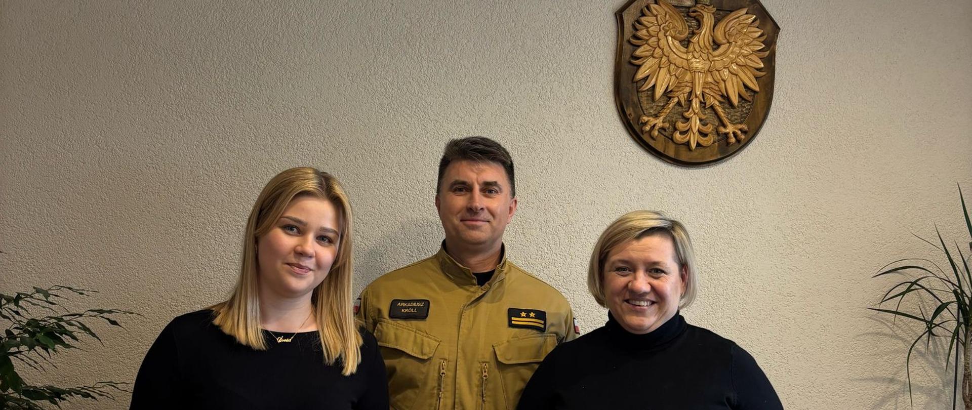 Trzy osoby stoją obok siebie. Z lewej strony stoi kobieta. Kobieta ma ubraną czarną bluzkę i niebieskie spodnie. Obok stoi Komendant Powiatowy PSP w Tucholi. Komendant ma na sobie mundur w kolorze piaskowym. Po prawej stronie stoi kobieta. Kobieta ma ubrany czarny golf i niebieskie spodnie. Kobiety na zdjęciu trzymają niebieskie teczki z logo Państwowej Straży Pożarnej. Na teczkach widnieje napis: ,,Komenda Powiatowa Państwowej Straży Pożarnej w Tucholi”. W tle na szarej ścianie widać drewniane godło Polski. Po bokach widać liście kwiatów. 