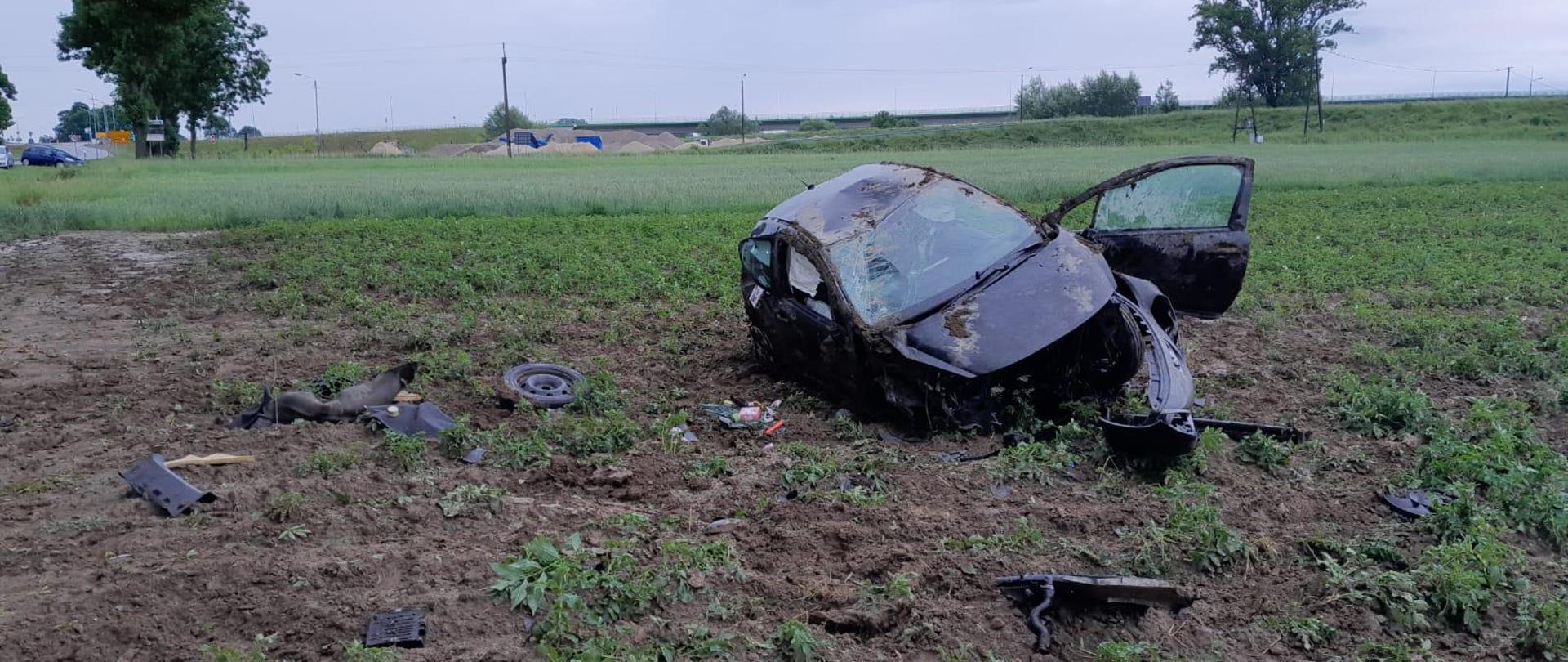 Rozbity samochód na polu, wokół niego leżą części od samochodu, w tle linki prądowe, słupy i kupki piasku. 