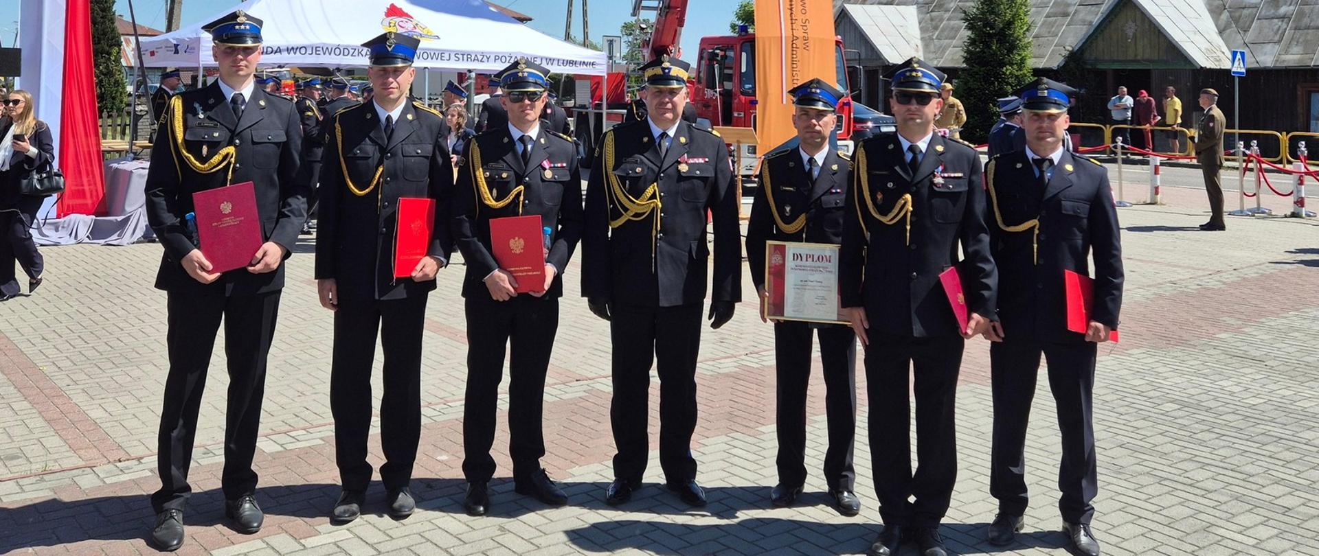 Obraz jest poziomym zdjęciem plenerowym, wykonanym przy jasnym świetle słonecznym w Polsce. Przedstawia oficjalną ceremonię z udziałem strażaków, stojących przed dużą drabiną strażacką i namiotami imprezowymi.