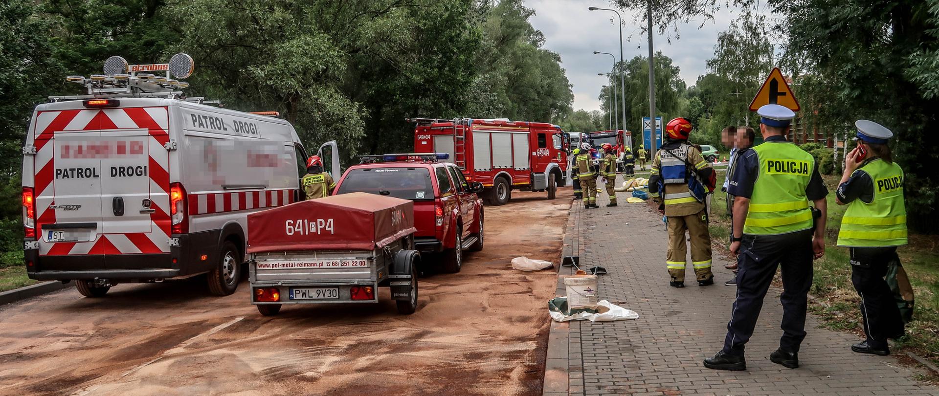 Jezdnia zasypana sorbentem. Na jezdni stoją wozy strażackie, na chodniku policjanci i strażacy.