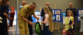 Strażak wręcza plecak dziewczynce obok stoją policjanci mężczyzna oraz dziewczynka w żółtej kamizelce która trzyma nagrody. 