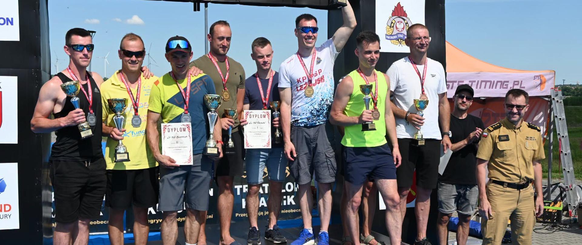 Zdjęcie przedstawia zawodników po dekoracji medalami, po wręczeniu pucharów i dyplomów. Na każdym stopniu podium stoi trzech zawodników – tylu z ilu składa się sztafeta. Każdy z zawodników trzyma w ręku puchar, każdy ma na szyi zawieszony pamiątkowy medal. Ponadto w rękach kilku zawodników znajdują się pamiątkowe dyplomy. Po lewej stronie na podium stoi 3 zawodników reprezentujących Komendę Powiatową Państwowej Straży Pożarnej w Bochni. Zawodnicy stoją na czarnym podeście, za nimi widoczna czarna sceneria za podium. Po prawej stronie obok podium stoi funkcjonariusz Państwowej Straży Pożarnej, który wcześniej wręczał medale, puchary i dyplomy. Strażak ten jest w ubraniu służbowym w kolorze piaskowym. Za tym strażakiem widać mężczyznę w czarnym ubraniu oraz namiot stanowiący element zaplecza technicznego imprezy. 