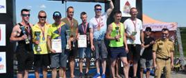 Zdjęcie przedstawia zawodników po dekoracji medalami, po wręczeniu pucharów i dyplomów. Na każdym stopniu podium stoi trzech zawodników – tylu z ilu składa się sztafeta. Każdy z zawodników trzyma w ręku puchar, każdy ma na szyi zawieszony pamiątkowy medal. Ponadto w rękach kilku zawodników znajdują się pamiątkowe dyplomy. Po lewej stronie na podium stoi 3 zawodników reprezentujących Komendę Powiatową Państwowej Straży Pożarnej w Bochni. Zawodnicy stoją na czarnym podeście, za nimi widoczna czarna sceneria za podium. Po prawej stronie obok podium stoi funkcjonariusz Państwowej Straży Pożarnej, który wcześniej wręczał medale, puchary i dyplomy. Strażak ten jest w ubraniu służbowym w kolorze piaskowym. Za tym strażakiem widać mężczyznę w czarnym ubraniu oraz namiot stanowiący element zaplecza technicznego imprezy. 
