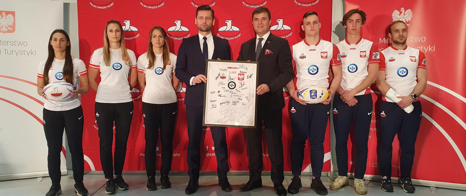 Polska zorganizuje Mistrzostwa Europy w rugby 7. Impreza została odebrana Rosji. Minister Kamil Bortniczuk i prezes Polskiego Związku Rugby Dariusz Olszewski w otoczeniu zawodniczek i zawodników reprezentacji Polski w rugby 7. Minister trzyma pamiątkową koszulkę z podpisami graczy.