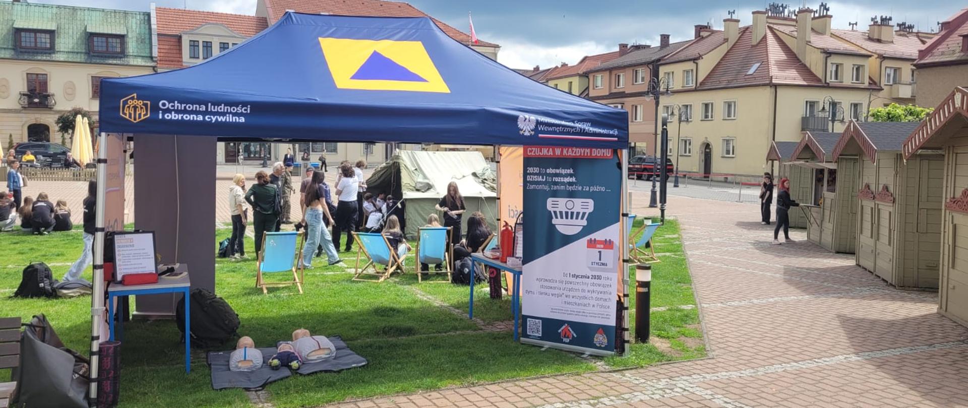 Widoczny namiot z niebieskim dachem i znakiem obrony cywilnej na środku. w namiocie stoją dwa stoliki, obok jednego z nich oparty plecak, na podłodze leżą fantomy szkoleniowe z pierwszej pomocy. Na namiocie napis: ochrona ludności i obrona cywilna, obok rol-up z czujką pożarową i napis czujka w każdym domu. W tle za namiotem spacerujący ludzie, miejskie kamienice i drewniane domki handlowe. 