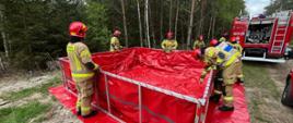 Strażacy rozstawiają zbiornik na wodę w lesie. Obok stoi samochód pożarniczy.