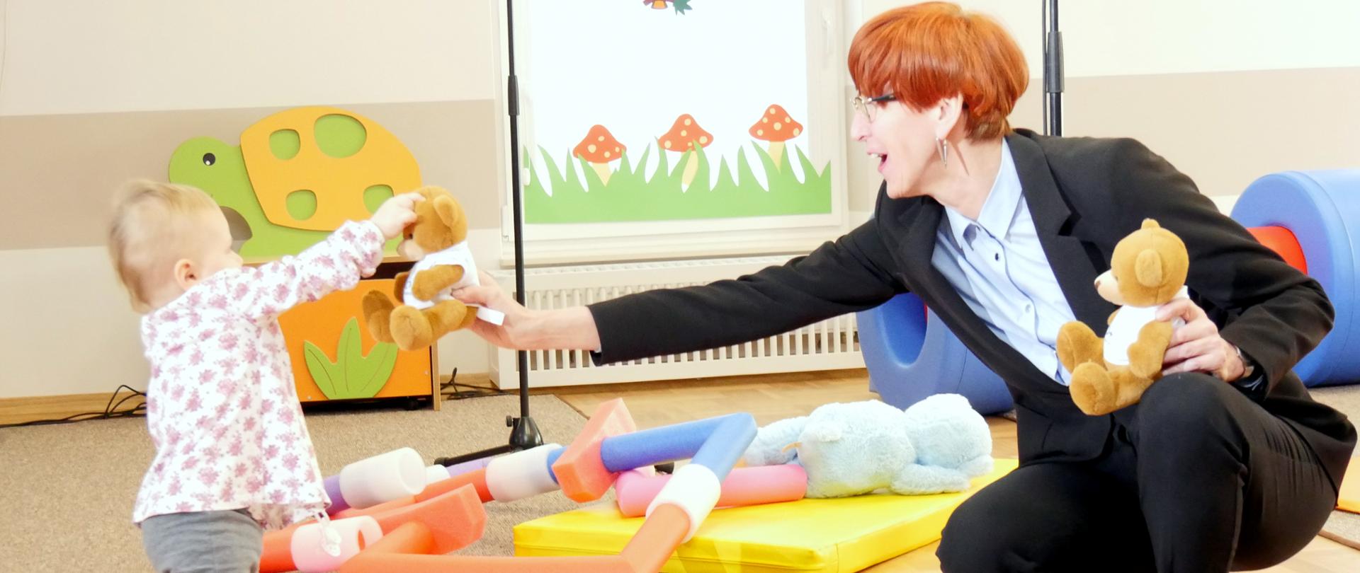 Otwarcie żłobka w Stalowej Woli. Minister rodziny Elżbieta Rafalska kuca i podaje misia pluszowego małemu dziecku.