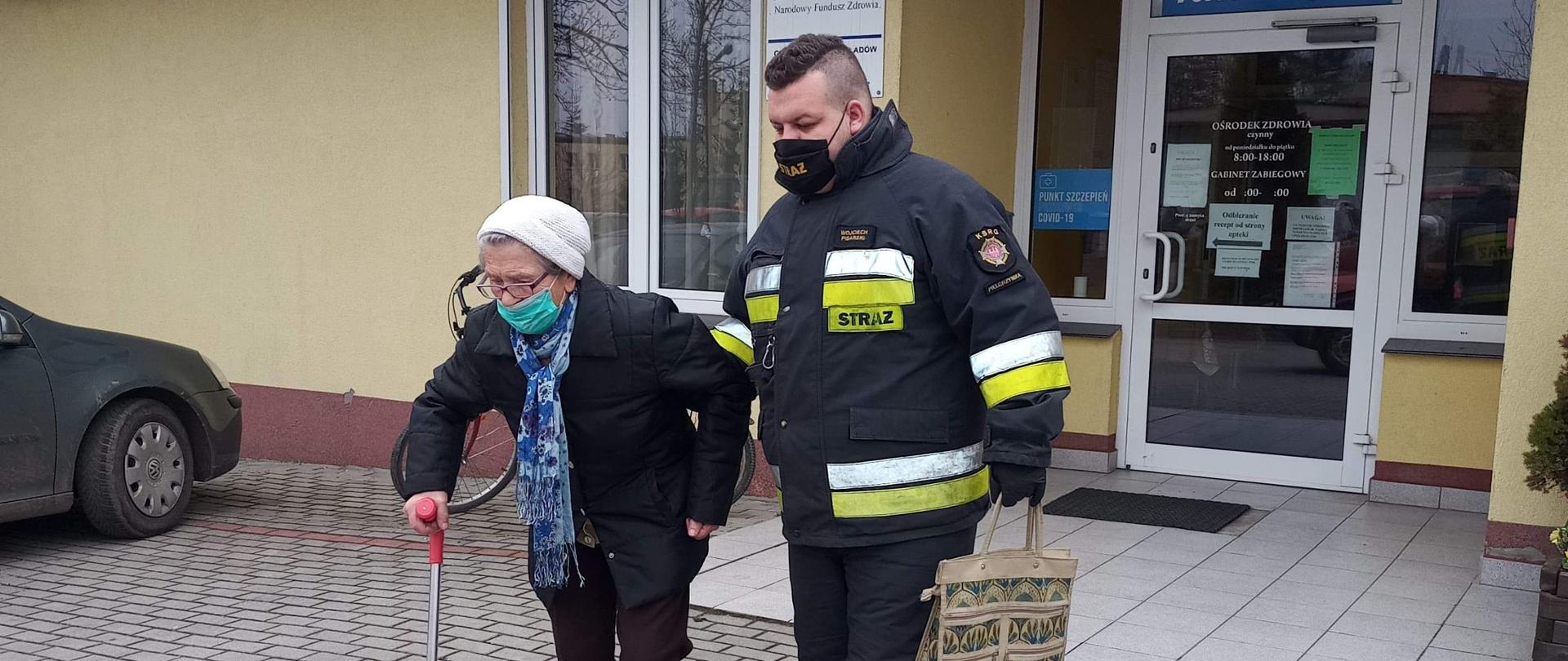 umundurowany strażak prowadzący starszą kobietę z budynku ośrodka zdrowia