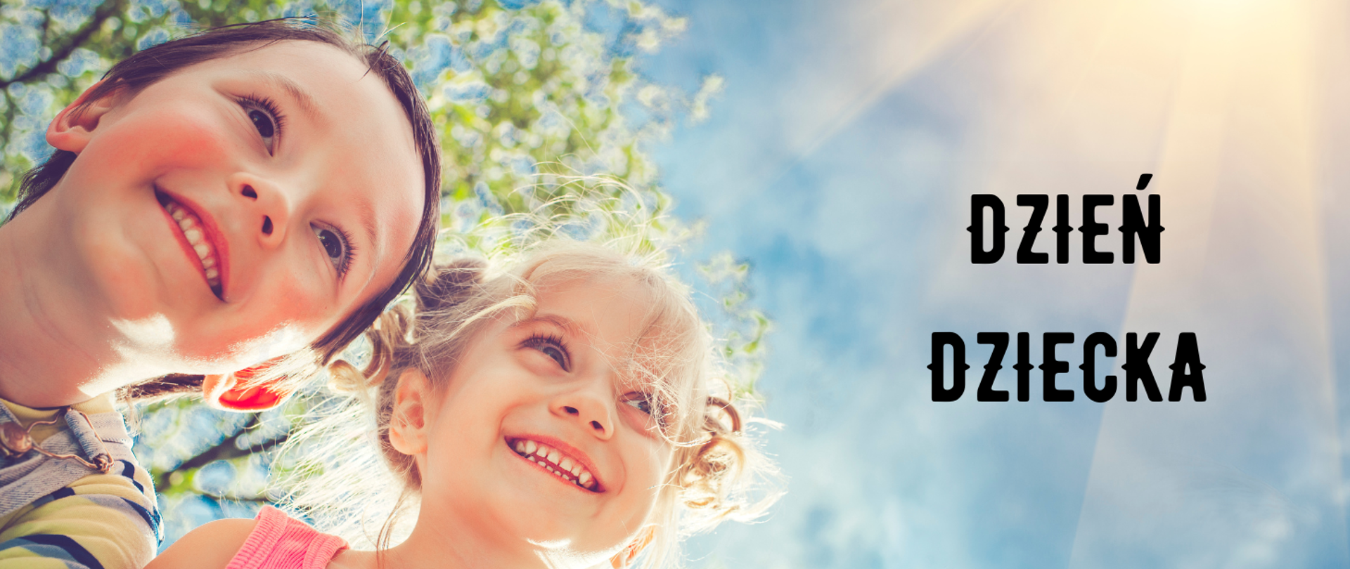 Po lewej stronie na zdjęciu dwie uśmiechnięte buzie dzieci. W tle za nimi rozmyte gałęzie drzewa oraz promienie słoneczne. Po prawej stronie czarny napis: "dzień dziecka".
