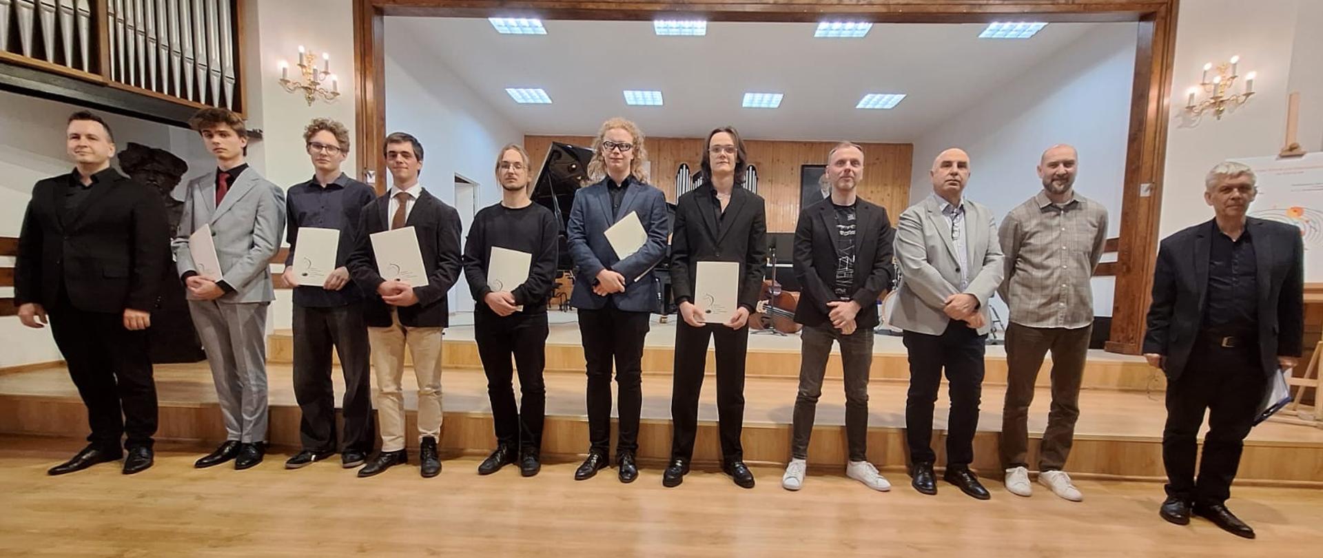 Grupa jedenastu mężczyzn — laureatów i jurorów konkursu improwizacji jazzowej — stoi w rzędzie na scenie sali koncertowej, trzymając pamiątkowe dyplomy.