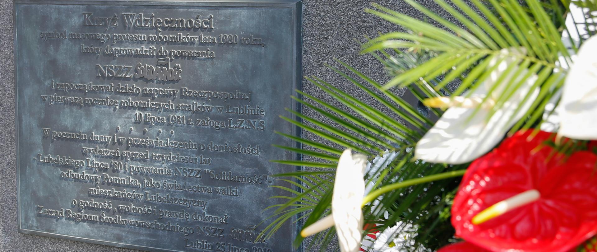 Tablica na Krzyżu Wdzięczności. Po prawej fragment biało- czerwonych kwiatów.