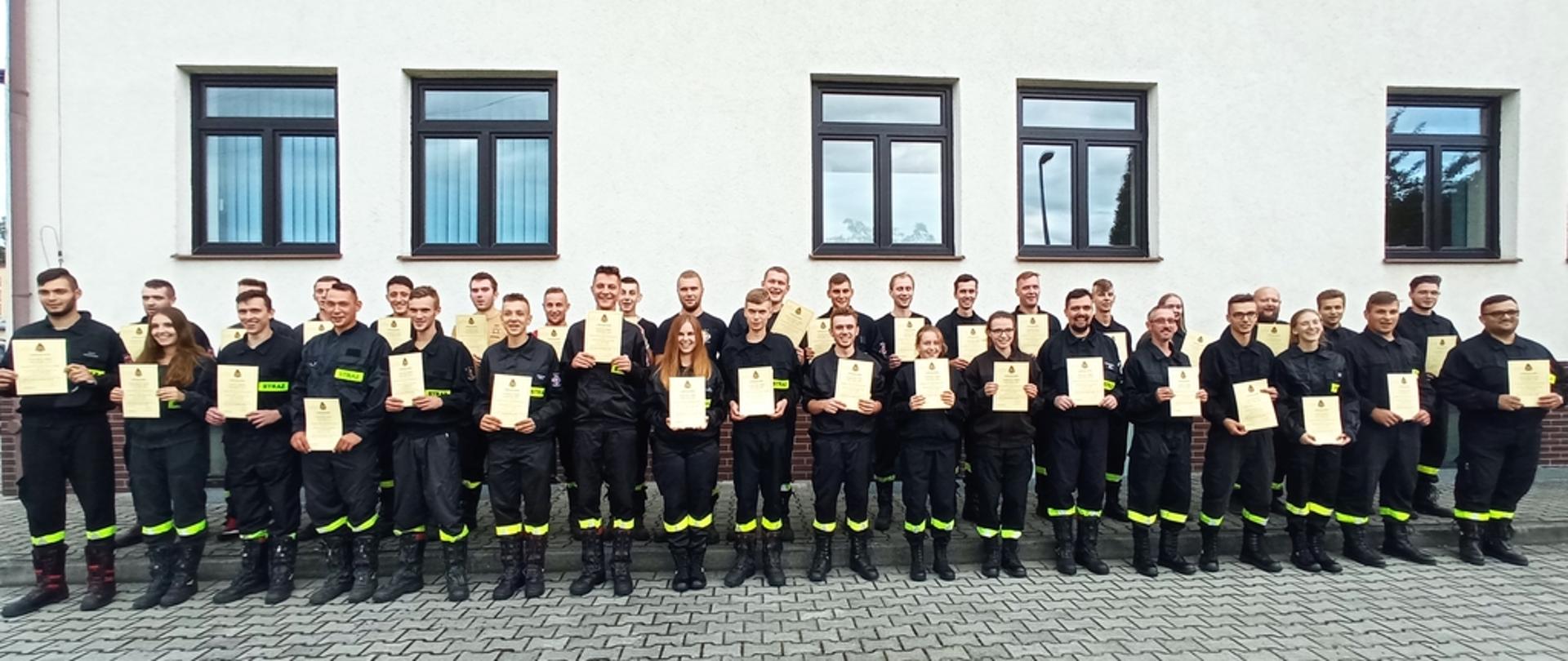 Na zdjęciu strażacy z Ochotniczych Straży Pożarnych po egzaminie kończącym szkolenie podstawowe strażaków ratowników OSP. Strażacy ubrani w ubrania koszarowe koloru czarnego bez czapek.