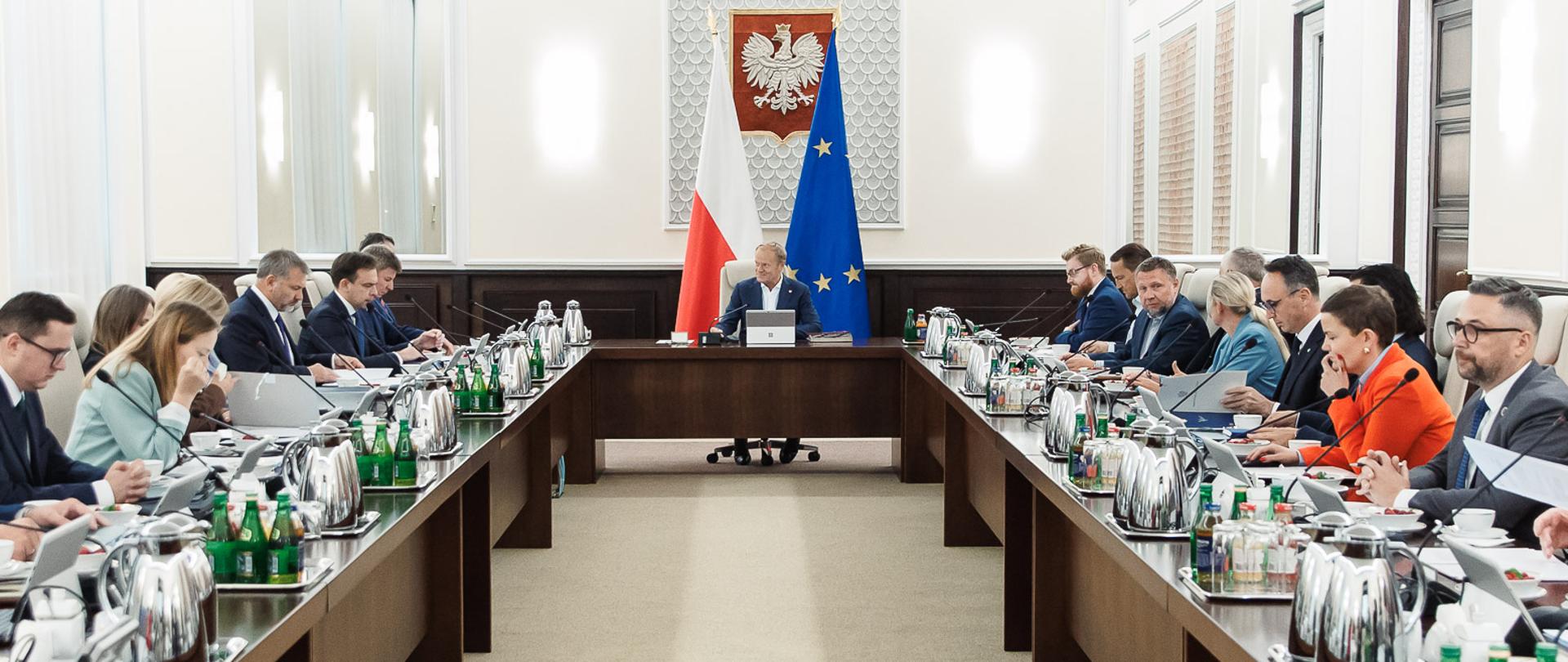 Posiedzenie Rady Ministrów 26.08.2025 r. Premier Donald Tusk na pierwszym planie, po lewej i prawej stronie ministrowie. Za plecami Premiera flagi Polski i Unii Europejskiej.